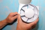 Hands holding a white exhaust fan against a blue wall; ready for installation.