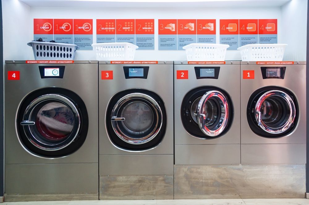Four stainless steel washing machines in a laundromat, with baskets and numbered labels.