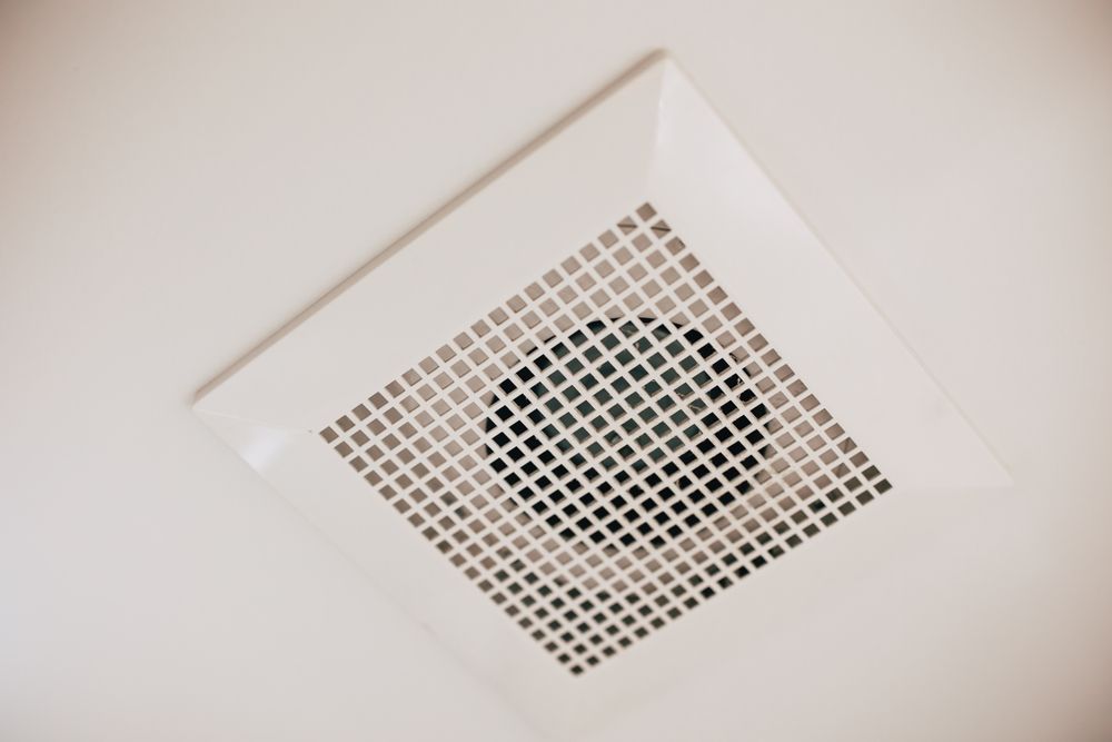 White ceiling-mounted bathroom vent with square grid and center fan, angled view.