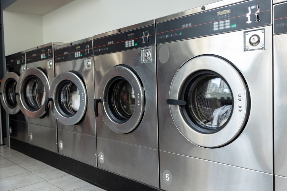 Row of silver washing machines in a laundromat, with black control panels and glass doors.