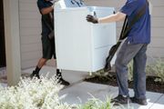Two people lifting a white washing machine outdoors with moving straps.