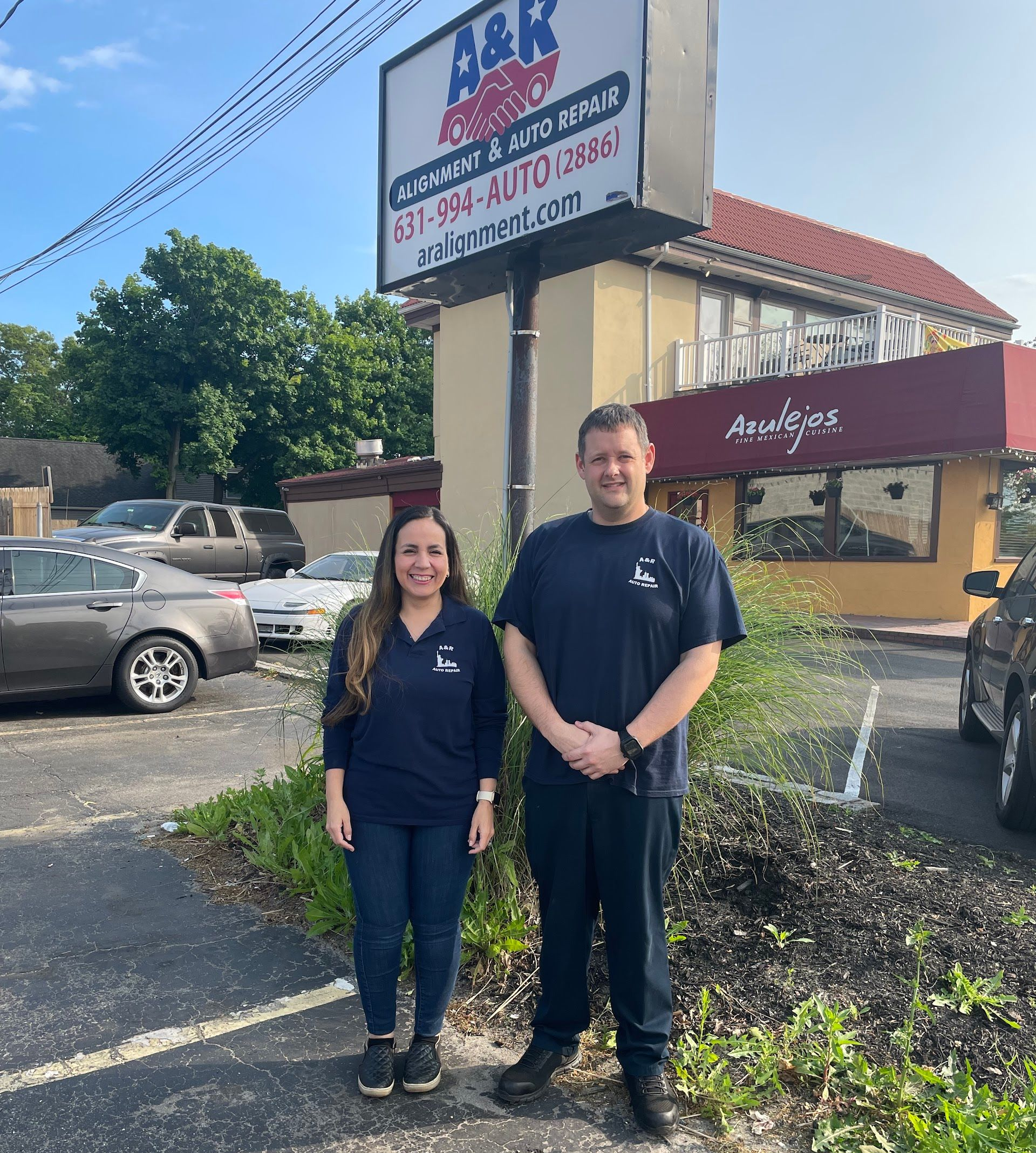 Two people, a woman and a man, in front of an auto repair shop sign, A&K, on a sunny | A & R Auto Repairday.