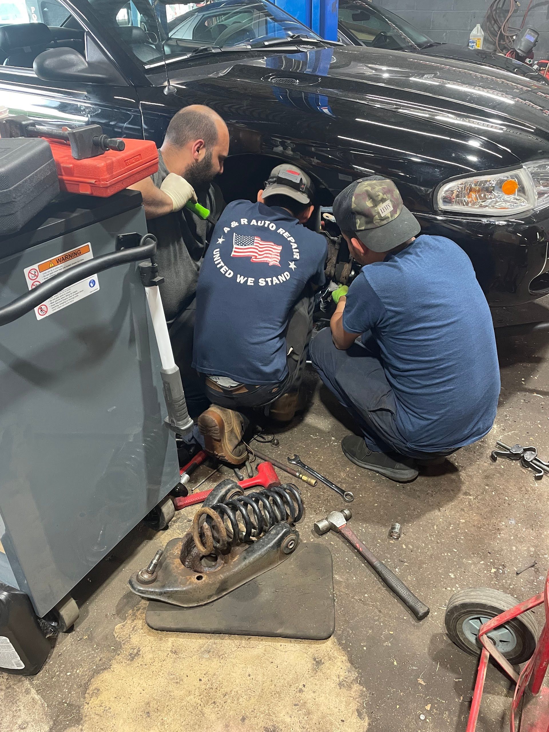 Three mechanics working on a black car tire at a garage. One holds a tool, two are crouched. Tools are strewn about. | A & R Auto Repair | A & R Auto Repair