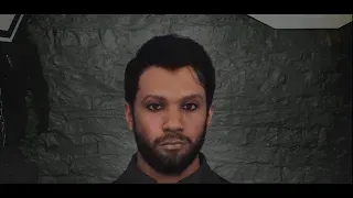 A man with dark hair and a beard stares intently at the camera. He's wearing a dark shirt and the background is a dark brick wall.