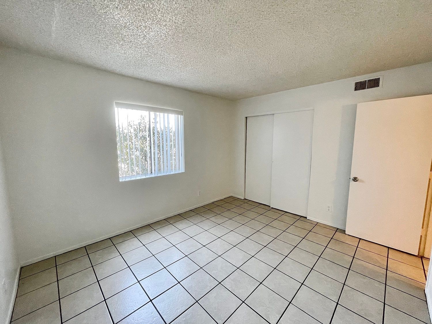 Empty room with white walls, tiled floor, sliding closet doors, a window, and a closed door.