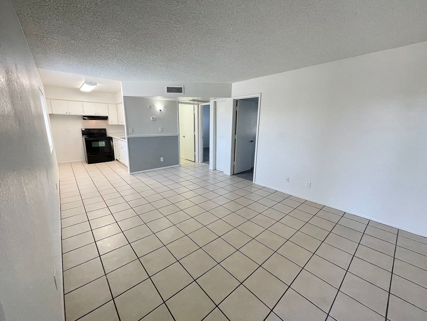 Empty, tiled-floor apartment interior. Kitchen and doorways visible. White walls, grey accent wall.