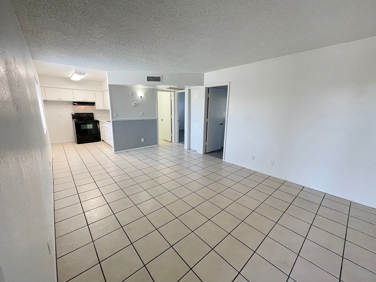 Empty, tiled-floor apartment interior. Kitchen and doorways visible. White walls, grey accent wall.