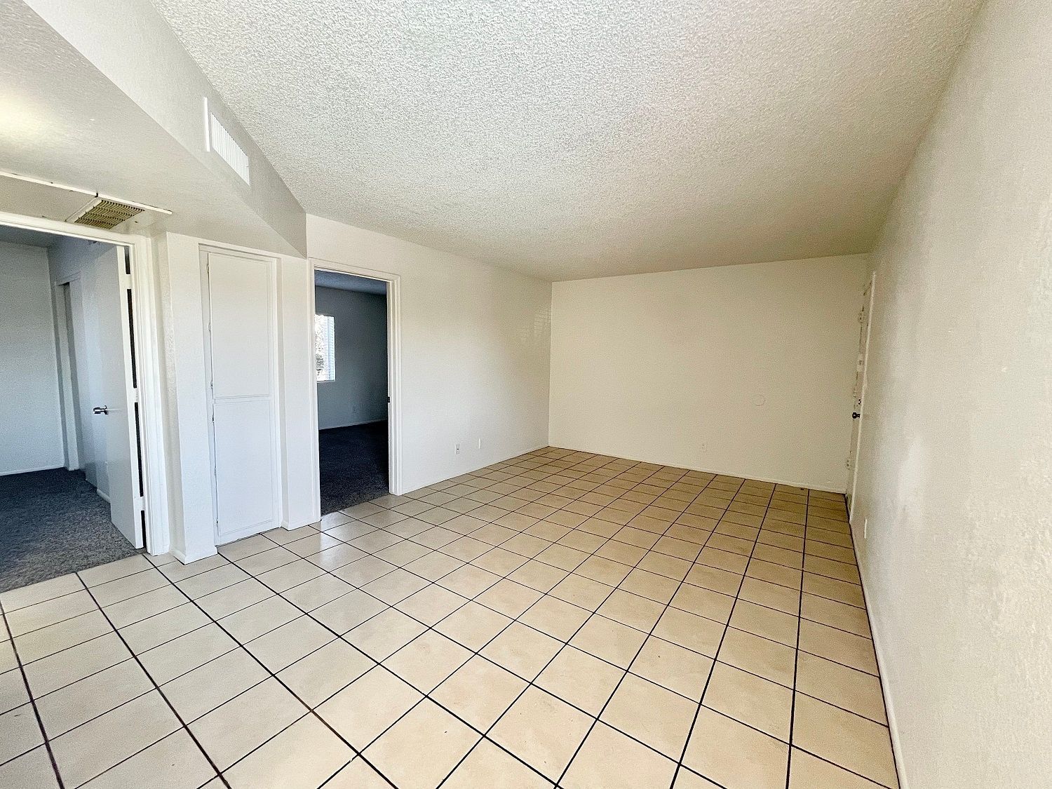 Empty, tiled-floor room with white walls, ceiling, and doors. A hallway and additional doorway are visible.