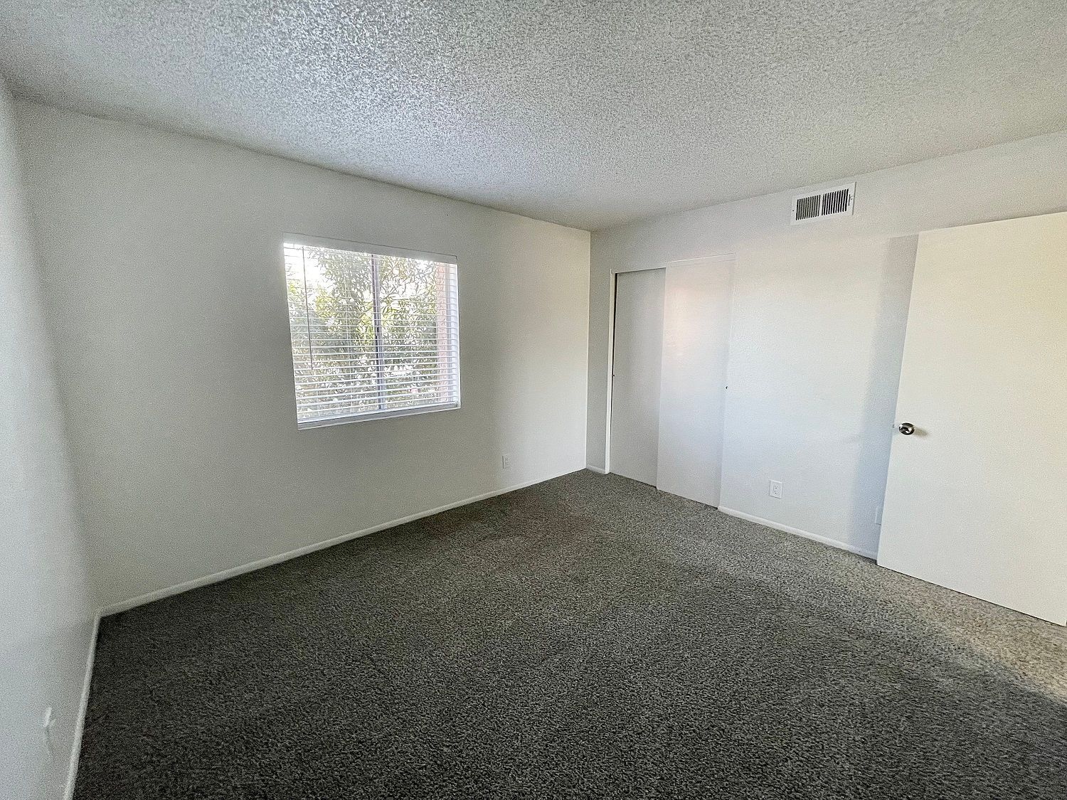 Empty room with dark gray carpet, white walls, window, door, and textured ceiling.