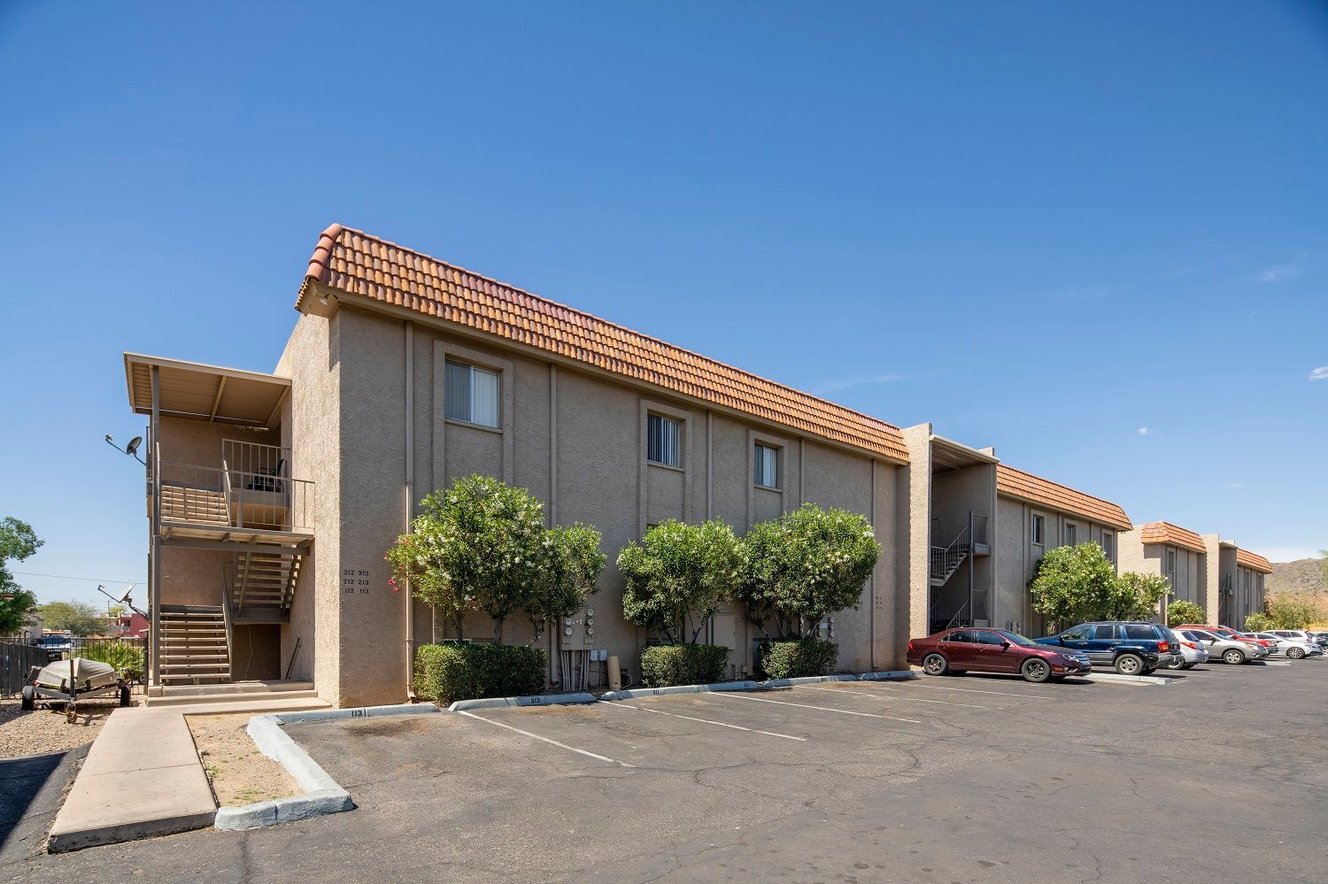 Apartment complex with tan walls, brown tile roof, and parked cars under a blue sky.