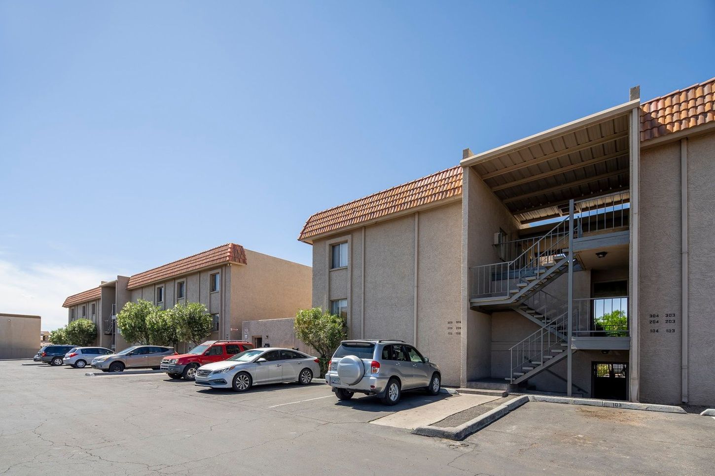 Apartment complex with multiple beige buildings, red tile roofs, and parked cars under a clear blue sky.