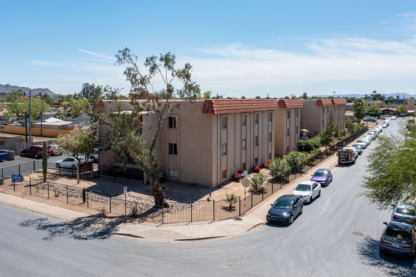 Apartment complex exterior on a sunny day; tan building with red tile roof, cars parked on street.