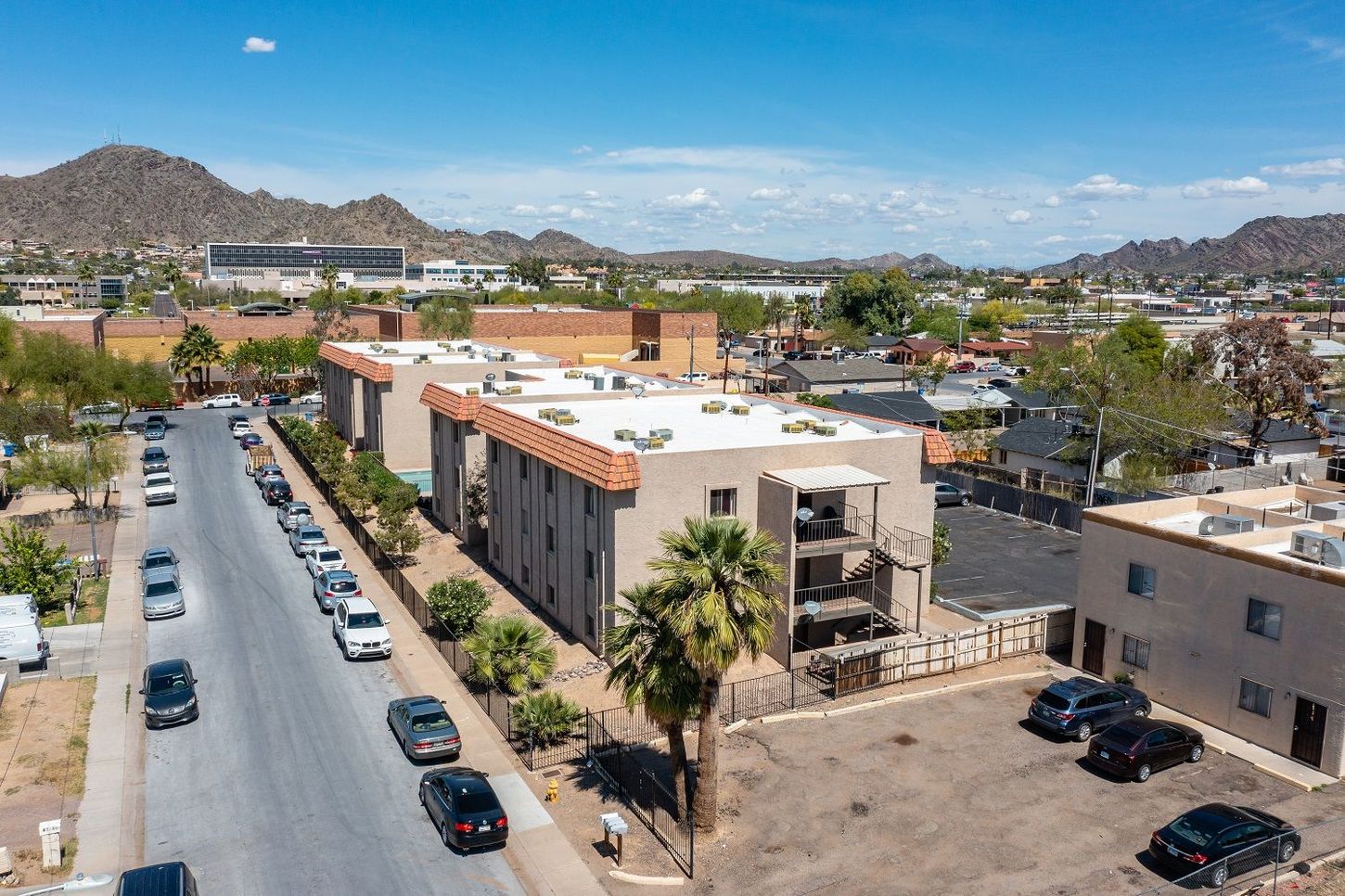 Aerial view of a street with parked cars, multi-story buildings, and mountains in the background under a blue sky.