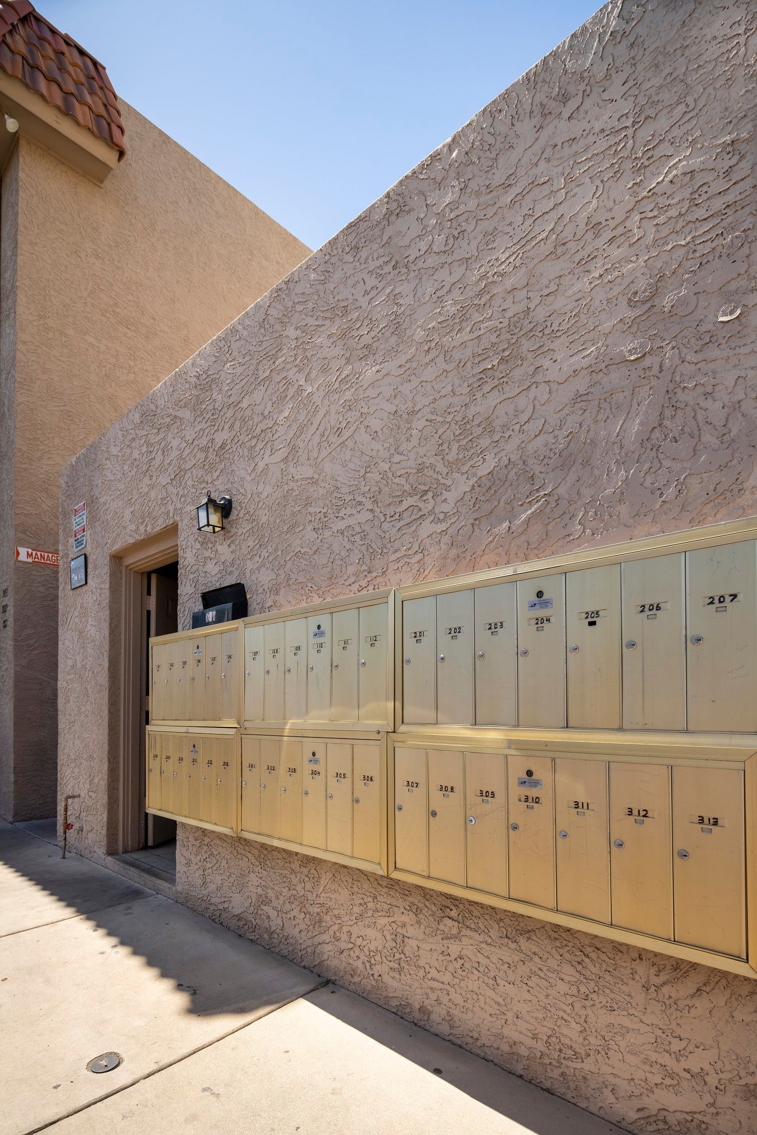 Mailboxes in a row on a tan stucco wall next to a sidewalk and a building entrance.