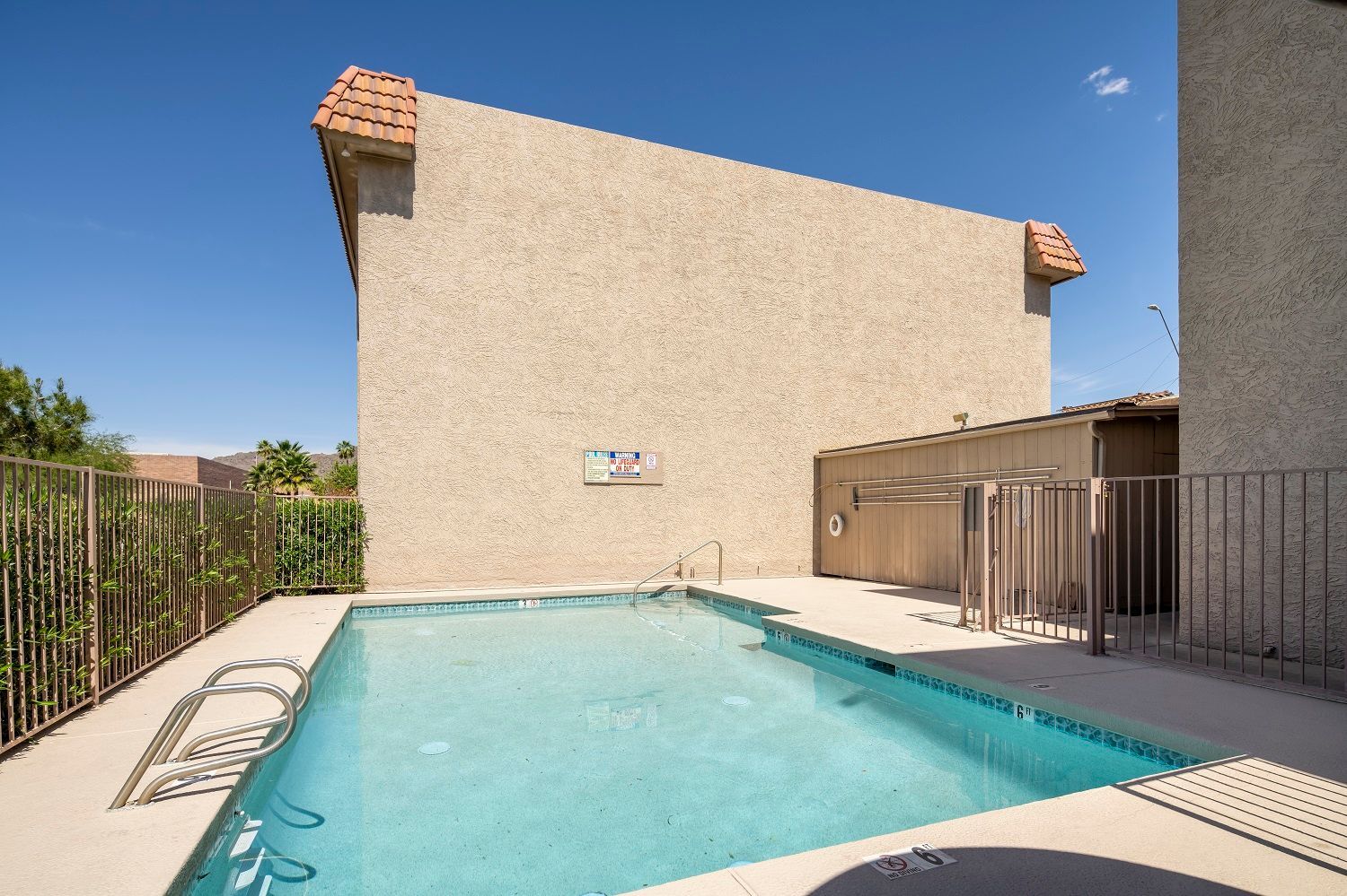 Swimming pool surrounded by a beige building and fence under a clear blue sky.