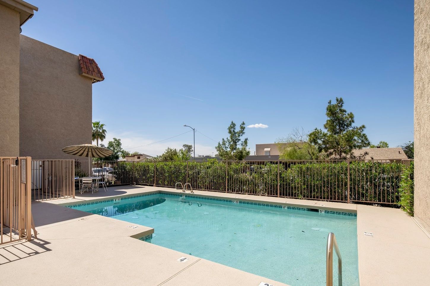 Swimming pool in a sunny outdoor area, beige deck, tan fence, green bushes, and a blue sky.