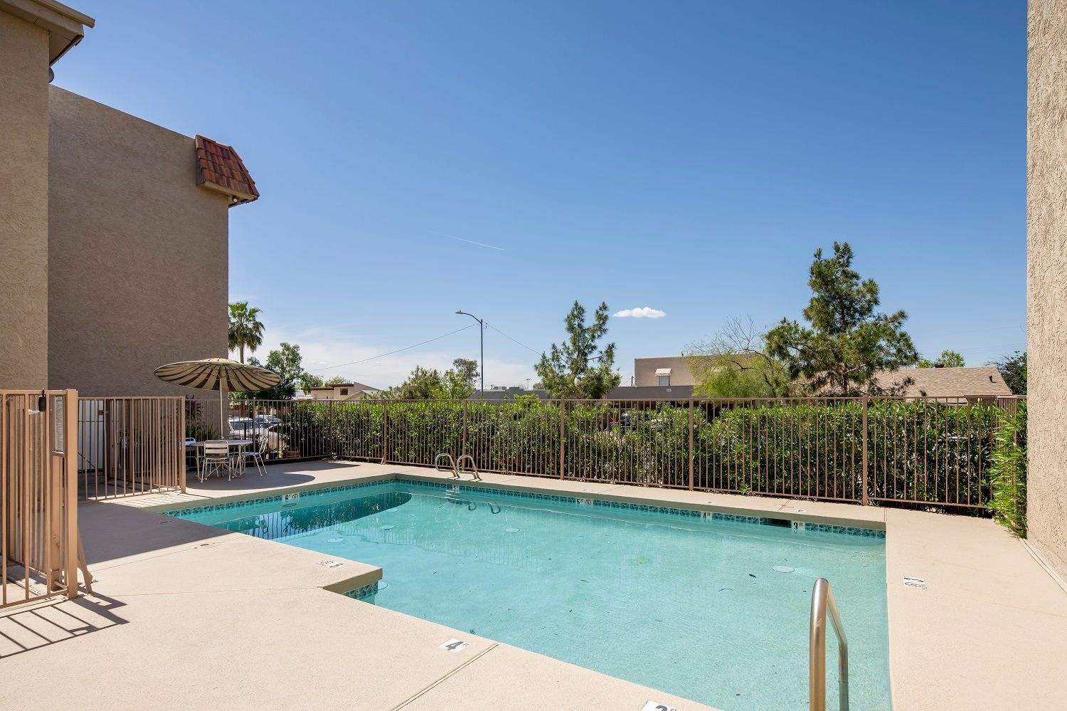 Swimming pool in a sunny outdoor area, beige deck, tan fence, green bushes, and a blue sky.