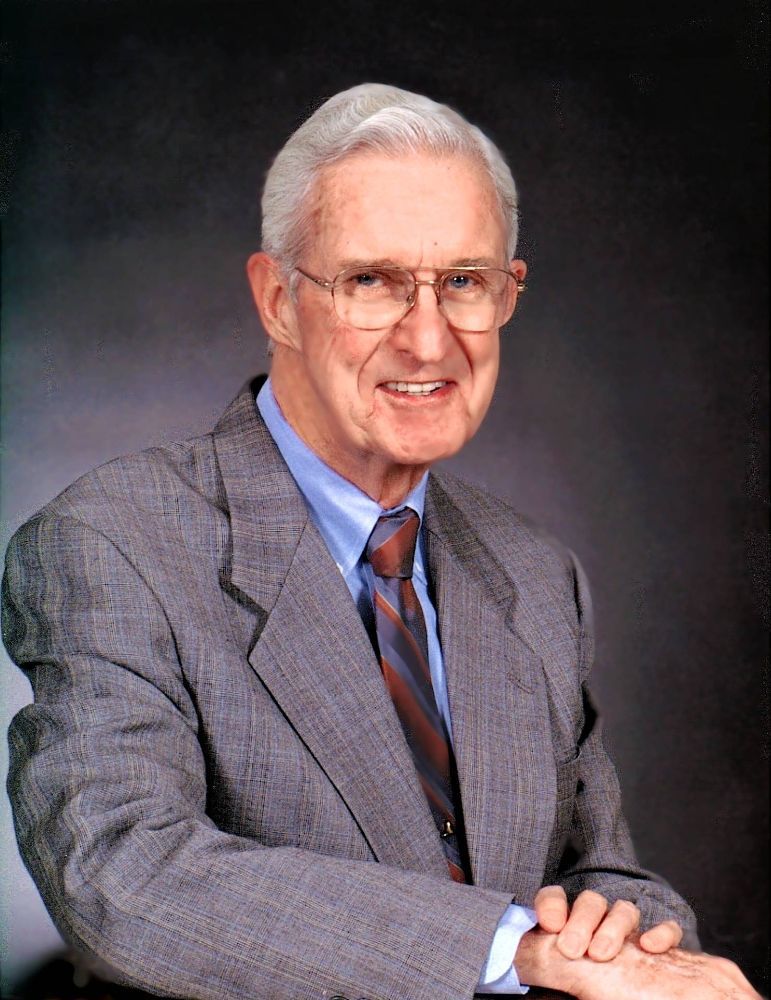 A person wearing a grey suit, blue dress shirt, and striped tie, smiling against a dark, textured background.