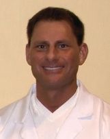 A headshot of a smiling person wearing a white medical lab coat against a plain, light-colored background.