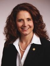 Headshot of a person with shoulder-length wavy brown hair, wearing a white collared shirt, pearl necklace, and blazer.