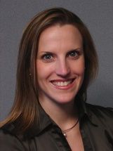 A smiling person with shoulder-length brown hair, wearing a dark olive-colored button-down shirt against a grey background.