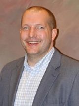 A smiling person with short hair, wearing a light blue and white checkered shirt under a grey blazer against a backdrop.