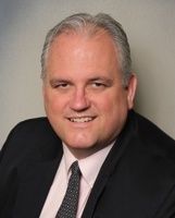A person in a dark suit, white collared shirt, and grey tie smiles against a neutral background.