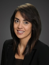 Headshot of a smiling person with shoulder-length brown hair, wearing a black blazer against a grey background.