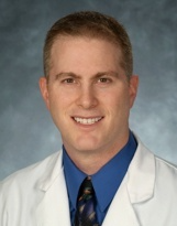 Headshot of a person wearing a white lab coat, blue dress shirt, and patterned tie against a mottled blue background.