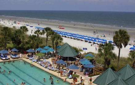 An elevated view of a hotel pool area with blue umbrellas, palm trees, and a sandy beach with beach chairs by the ocean.