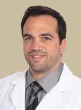 A person in a white medical coat, gray shirt, and patterned tie smiling against a neutral tan background.