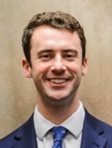 A person with short brown hair, wearing a navy suit, white shirt, and blue patterned tie, smiles against a tan background.