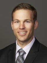 A headshot of a person smiling, wearing a dark suit, light shirt, and patterned tie against a neutral grey background.