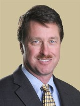 Headshot of a man with short brown hair and a mustache, wearing a suit and patterned tie against a plain tan background.