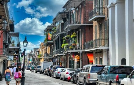 A street scene in New Orleans featuring historic brick buildings with iron balconies, parked cars, and pedestrians.