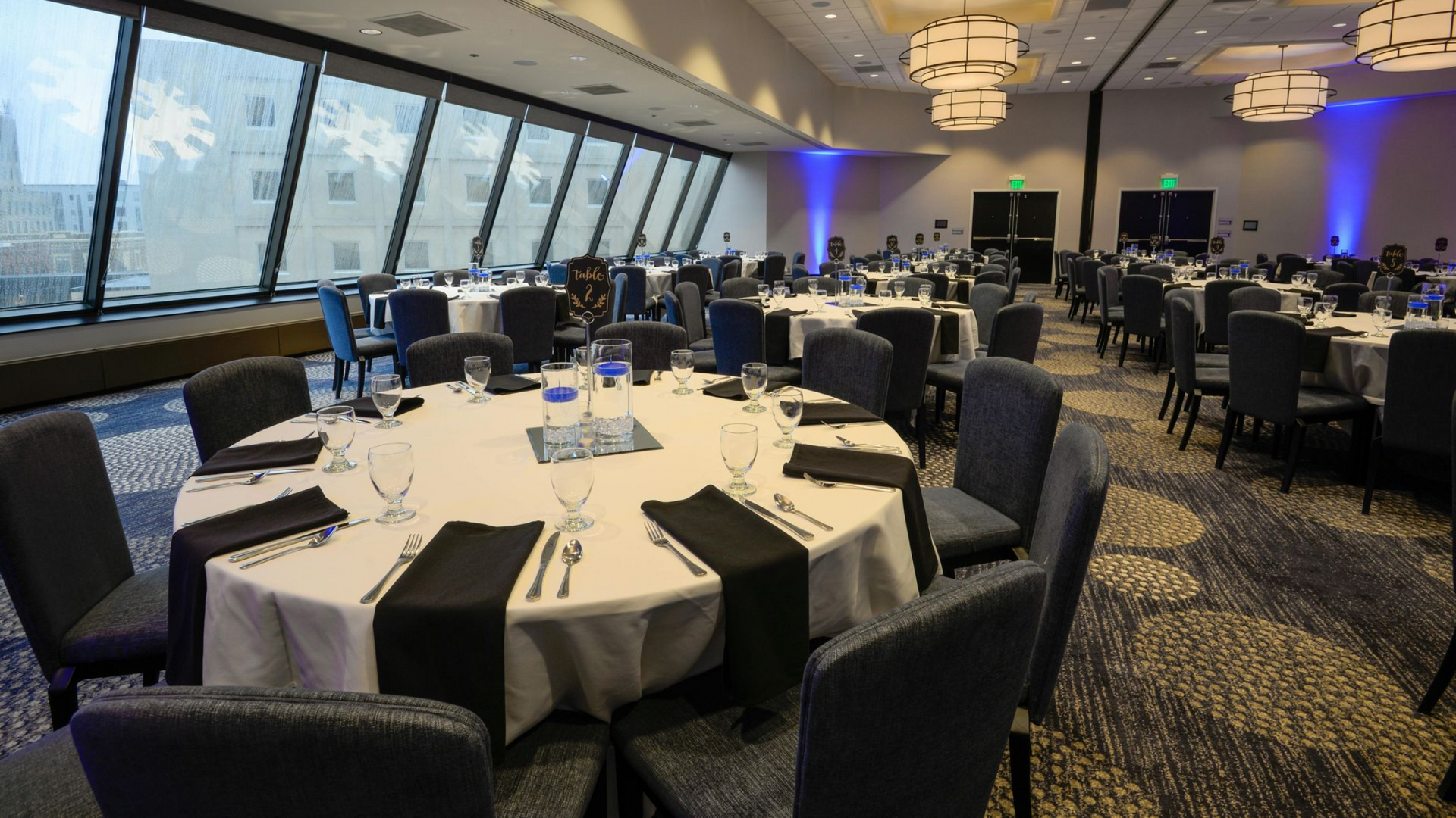 A modern banquet hall with round tables set for dining, dark chairs, large windows, and decorative ceiling lights.