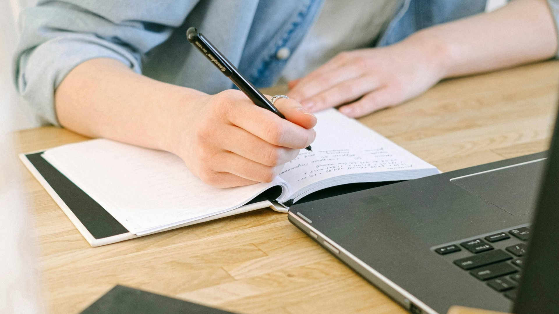 A person writing in a notebook next to a laptop on a wooden desk.