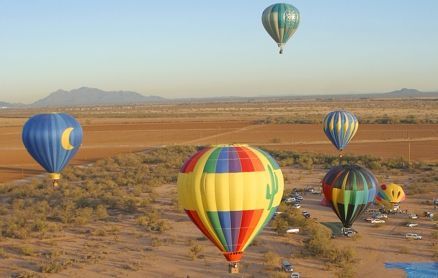 Several hot air balloons float over a desert landscape with scrub brush and distant mountains under a clear sky.