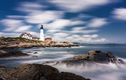 Portland Head Light in Maine, standing on a rocky coastline beneath a dramatic, long-exposure cloudy blue sky.