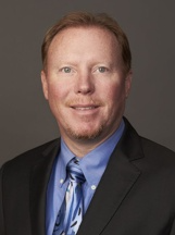 Headshot of a man with light brown hair wearing a dark suit, blue collared shirt, and a blue patterned tie.