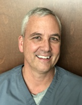 A person with short, graying hair wearing a blue scrub top and gray undershirt, smiling against a wood-toned background.