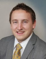 A person in a gray suit and patterned yellow tie smiles in a headshot against a blue and gray background.