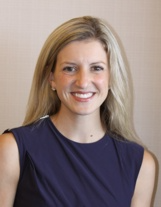 Headshot of a person with blonde hair wearing a navy blue sleeveless top, smiling against a neutral tan background.