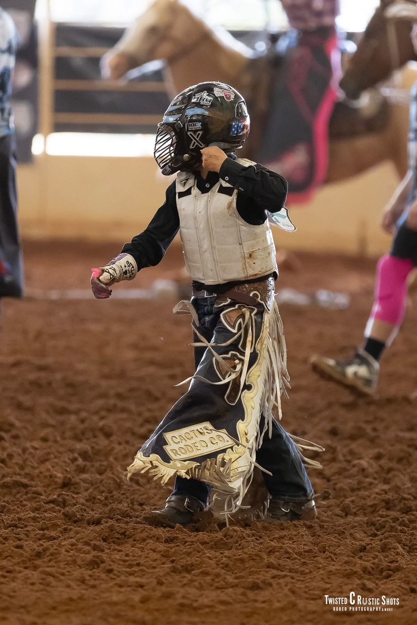 Young rodeo rider in safety gear, walking in a dirt arena.