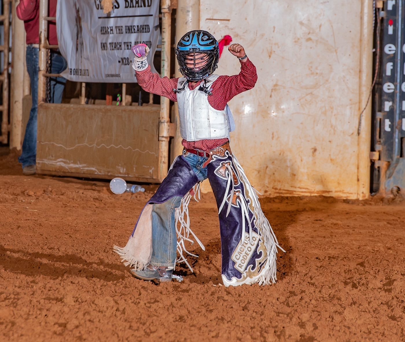 Young bull rider celebrating, wearing protective gear, in arena with red dirt.