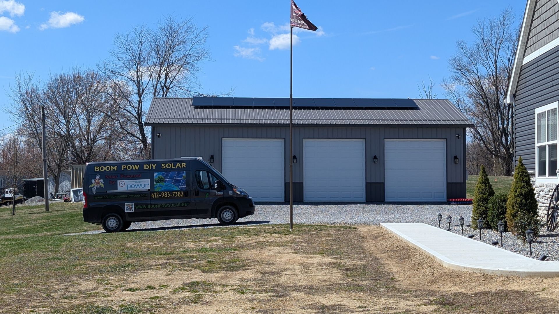 A gray van with a mural is parked in front of a three-door garage with solar panels on the roof, a flag, and a house on a sunny day.