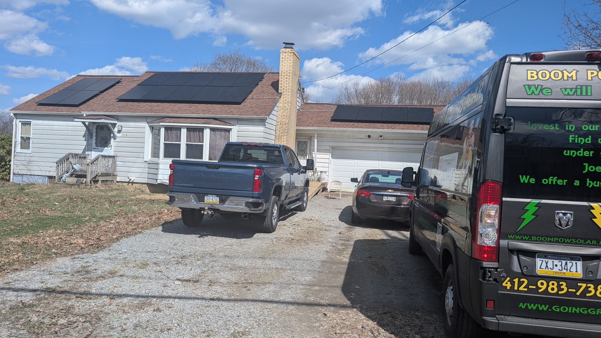 A rundown house with solar panels on the roof, a truck, a car, and a van parked in the driveway.