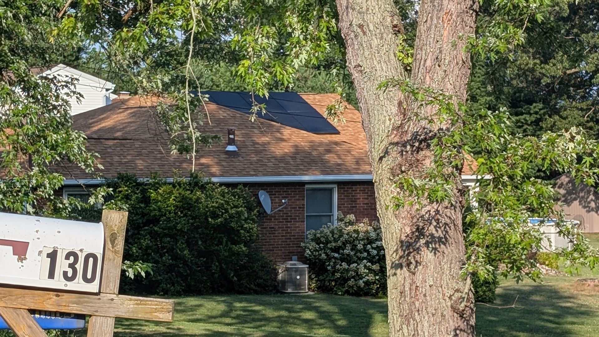 A house with a brick facade and brown roof behind a mailbox with the number 130, trees and bushes surrounding it.