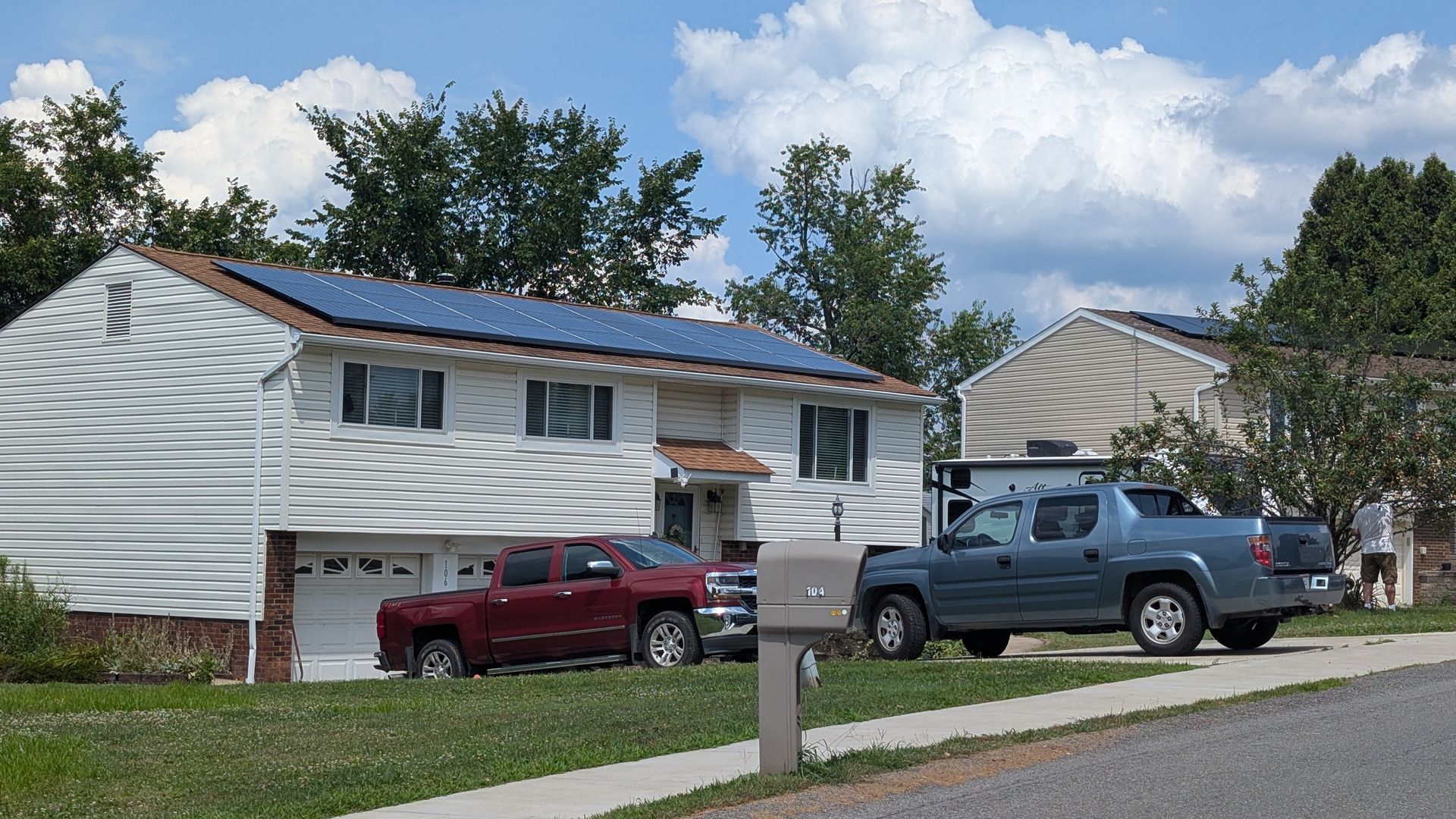 A suburban house with solar panels on its roof, a red pickup truck in the driveway, and a blue pickup truck parked on the street.