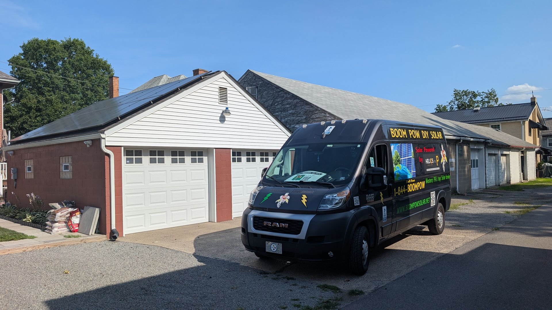 A black van with colorful logos parked in front of a garage with solar panels. The garage is red brick with a white door.